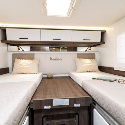 Twin single beds in a camper van with a central wood table, beige headboard, and white overhead cabinets showing a Benimar logo.