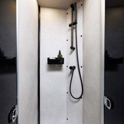 Shower stall interior with a handheld shower on the right wall above a small corner shelf holding a bottle, light fixtures, and a skylight above.