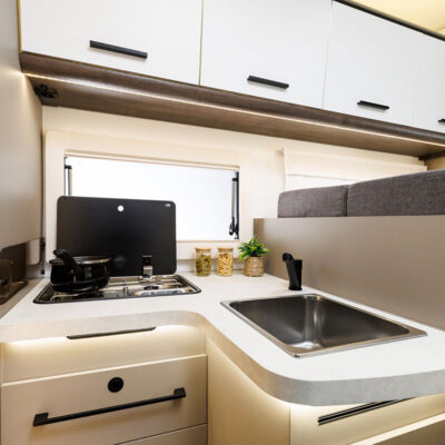 Compact camper van kitchen with a two-burner stove, sink, and overhead white cabinets; small window and decorative jars on the counter.