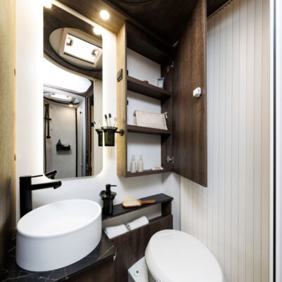 Compact bathroom with round white vessel sink, black faucet, and dark wood cabinetry; mirror and open shelves with toiletries above the sink.