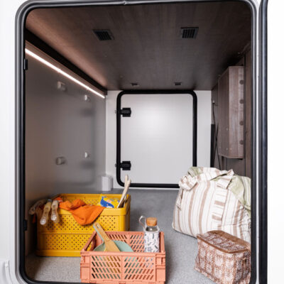 Open RV storage bay showing orange crates with tools, a striped bag, and a wicker-patterned chest on a gray carpeted floor with a wooden cabinet to the right.