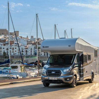 Silver motorhome parked at a marina with moored boats and white stone buildings in the background under a blue sky.
